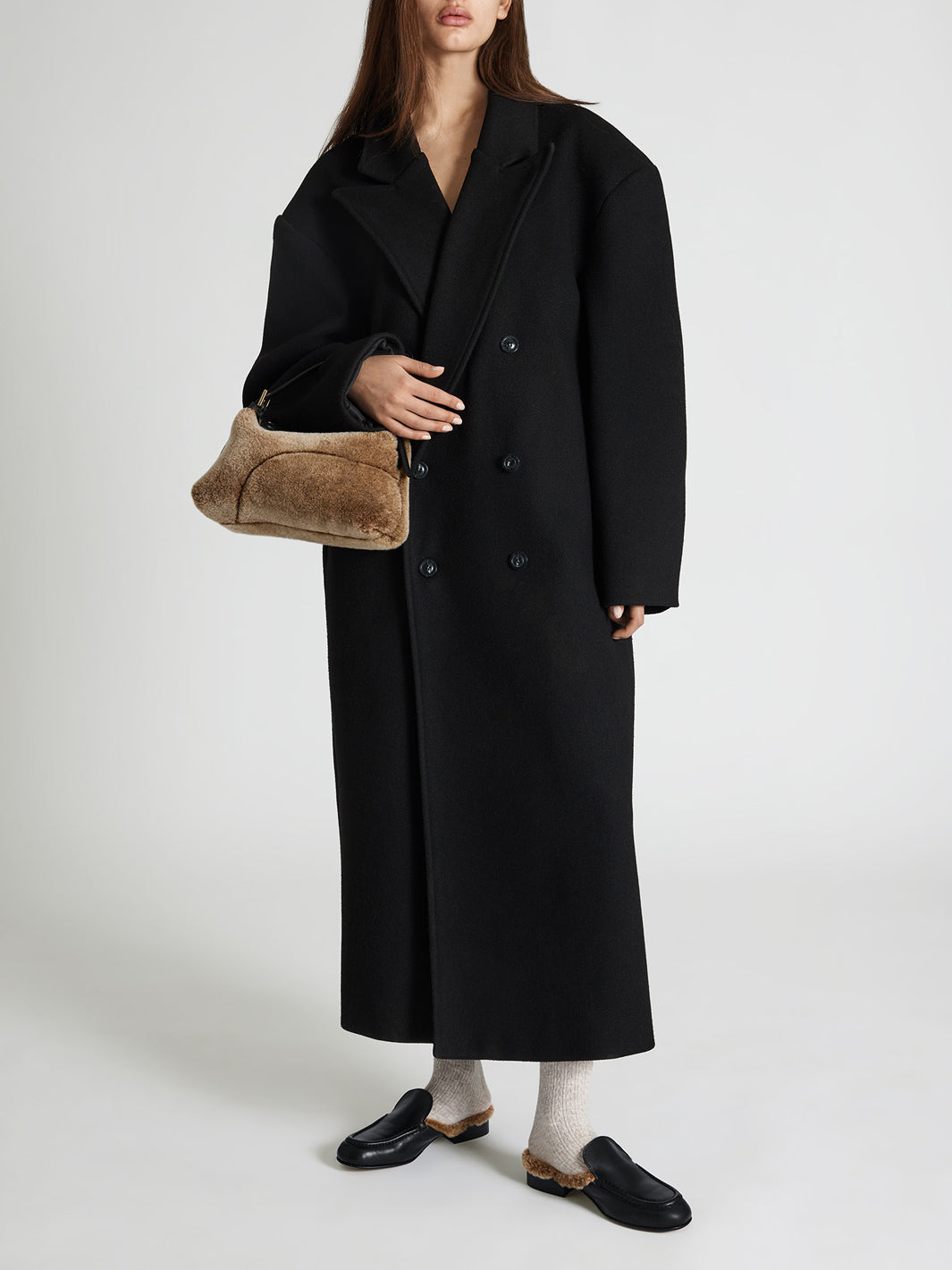 A model wearing a long black double-breasted coat, holding a brown textured hobo bag, paired with white socks and black loafers, against a light gray studio background.
