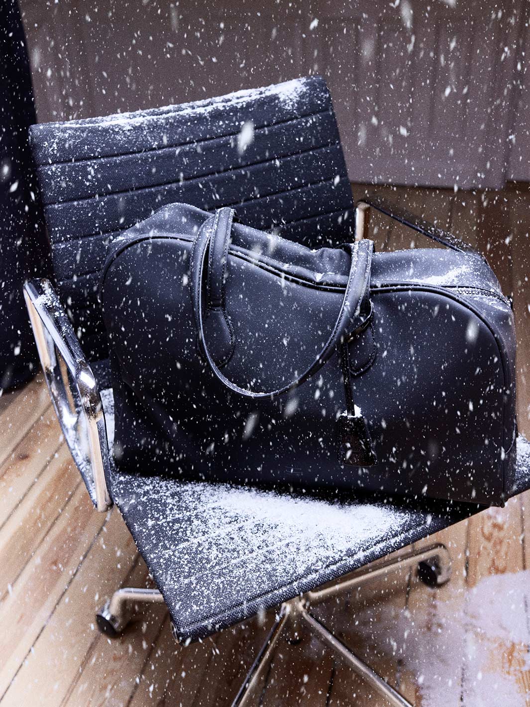 A black leather duffel bag with silver-toned hardware sits on a black office chair covered in white snow, with falling snow visible in the air.