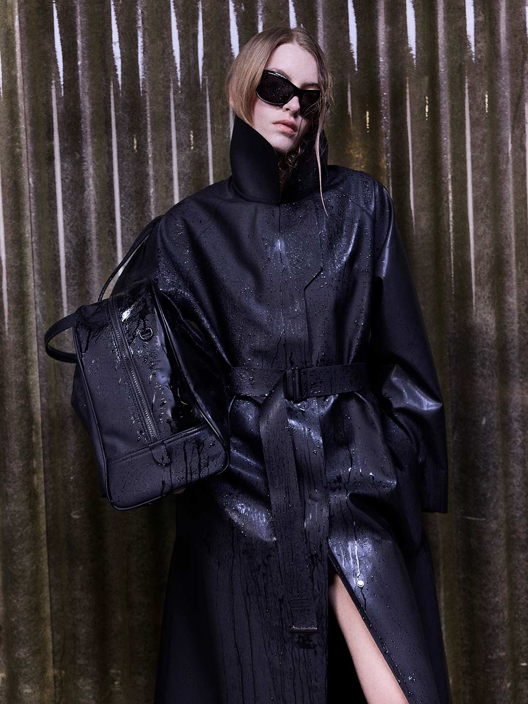 A model wearing a glossy black high-neck trench coat with a matching belt, black sunglasses, and carrying a black textured shoulder bag, posed against a dark corrugated metal curtain.