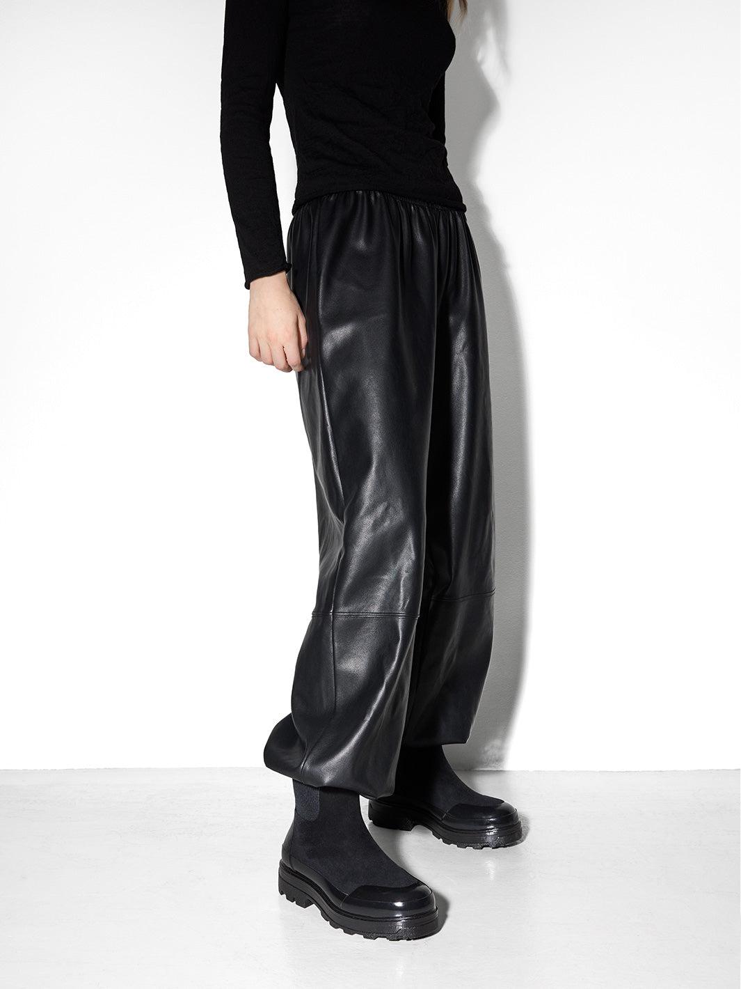 A model wearing a pair of black Chelsea-style ankle rain boots with a glossy patent leather toe cap and thick lug soles.