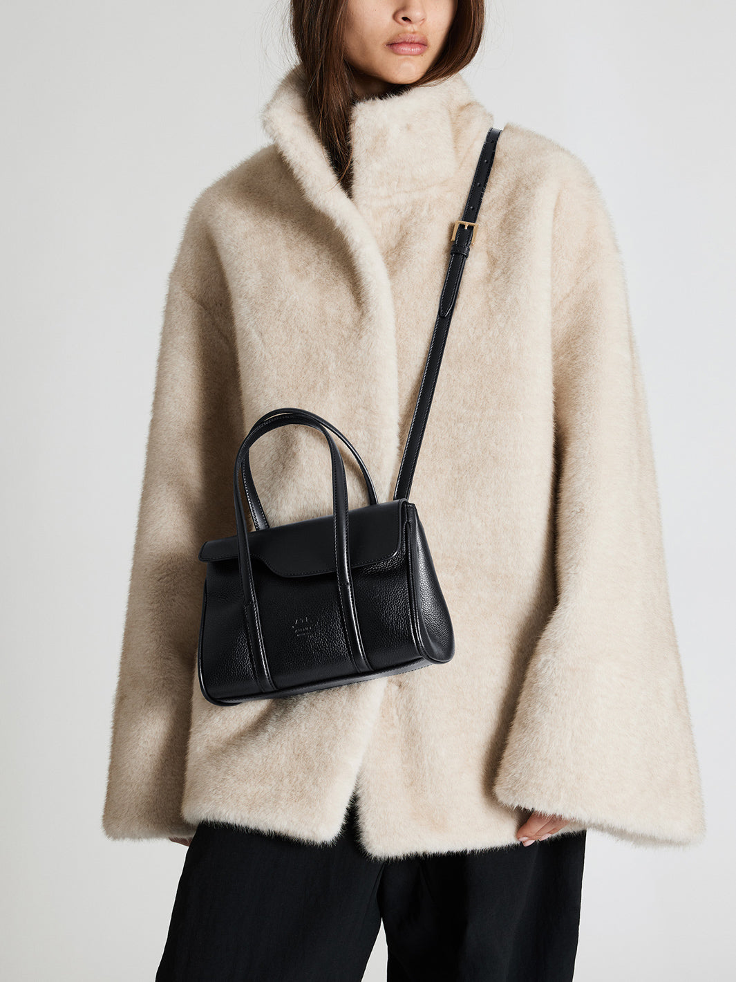 A model wearing a cream-colored fuzzy fur coat and black trousers, carrying a structured black leather handbag with a shoulder strap.