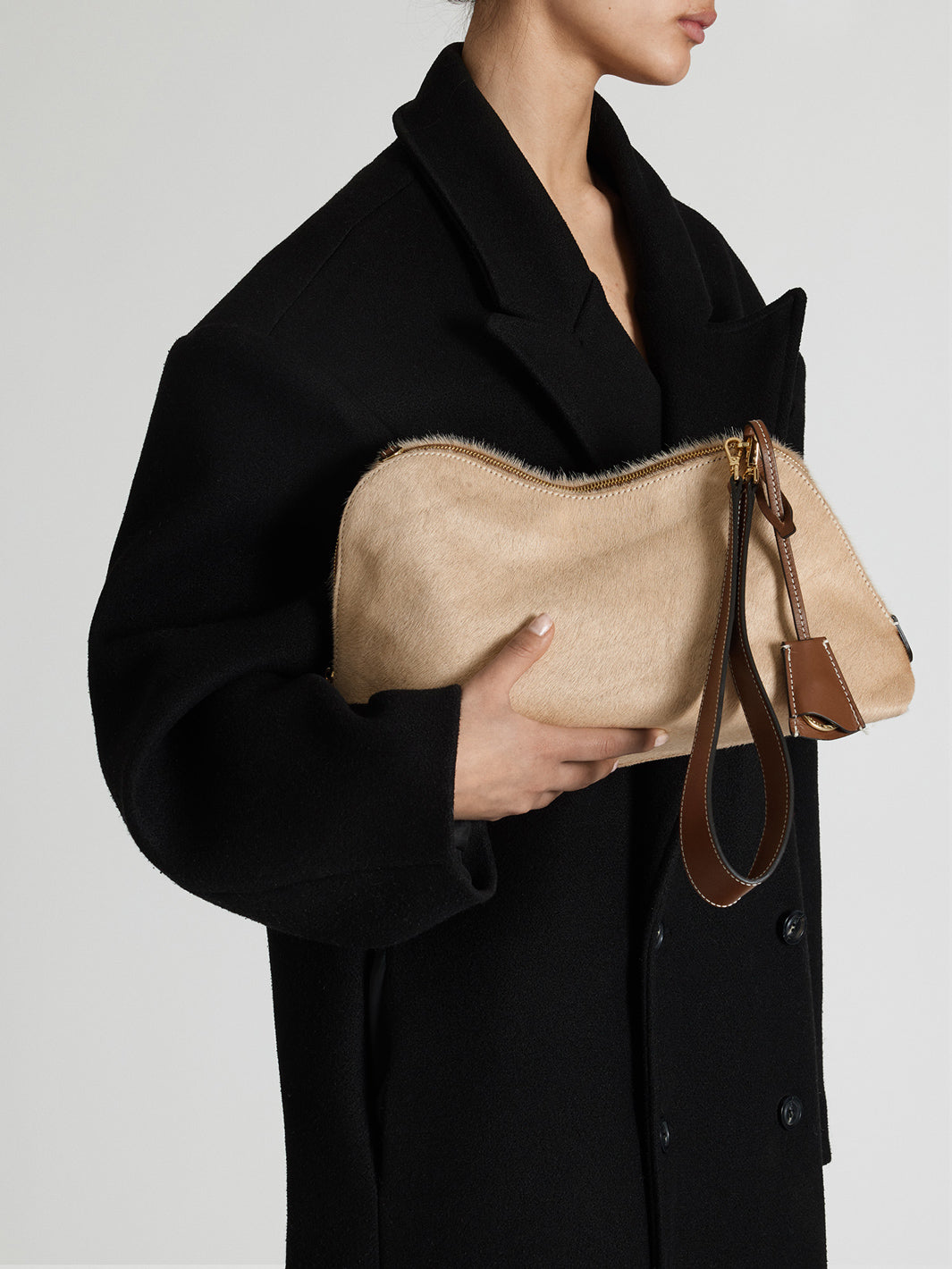 A model in a black coat holds a beige suede clutch bag with brown leather trim and a matching wristlet strap against a light gray background.