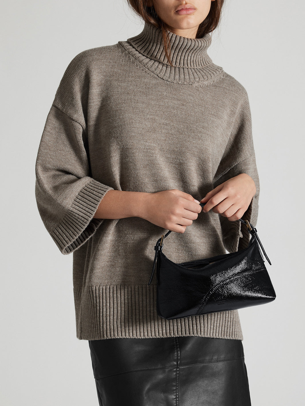 A model wearing an oversized ribbed turtleneck sweater and black leather skirt, holding a shiny black hobo handbag, photographed against a light gray studio background.